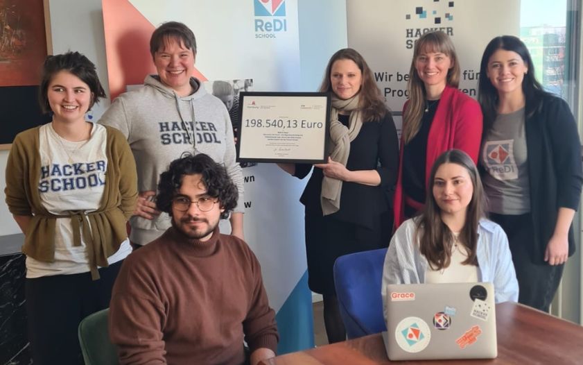 Eine Gruppe von sieben Personen, vier Frauen stehend und drei Personen sitzend, posiert gemeinsam für ein Foto in einem Raum mit Bannern der Organisationen
