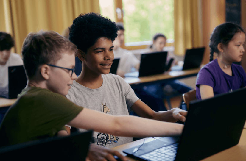 Schüler sitzen mit Laptops im Klassenraum und programmieren gemeinsam – im Vordergrund agieren zwei Jungs an einem Laptop und zeigen sich etwas.