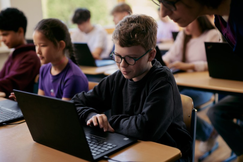 Ein Schüler mit Brille arbeitet an einem Laptop in einem Klassenzimmer mit anderen Schülern im Hintergrund.