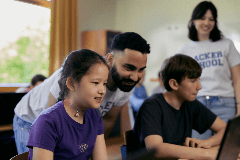 Zwei Kinder sitzen nebeneinander vor einem Laptop und schauen konzentriert auf den Bildschirm. Ein Mädchen im lila T-Shirt lächelt, ein Junge im dunkelblauen T-Shirt tippt auf der Tastatur. Hinter ihnen beugt sich ein junger Mann mit Bart zu ihnen und unterstützt sie freundlich. Im Hintergrund steht eine junge Frau und lächelt. Beide Erwachsenen tragen T-Shirts mit der Aufschrift