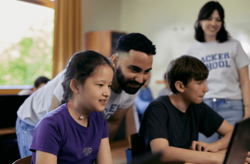 Zwei Kinder sitzen nebeneinander vor einem Laptop und schauen konzentriert auf den Bildschirm. Ein Mädchen im lila T-Shirt lächelt, ein Junge im dunkelblauen T-Shirt tippt auf der Tastatur. Hinter ihnen beugt sich ein junger Mann mit Bart zu ihnen und unterstützt sie freundlich. Im Hintergrund steht eine junge Frau und lächelt. Beide Erwachsenen tragen T-Shirts mit der Aufschrift