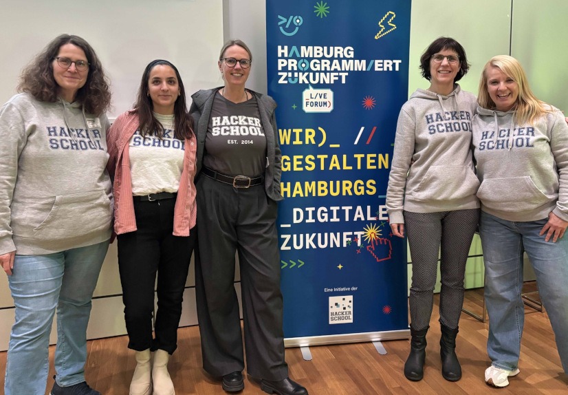 Fünf Mitarbeiterinnen der Hacker School neben einem Aufsteller des Projektes 